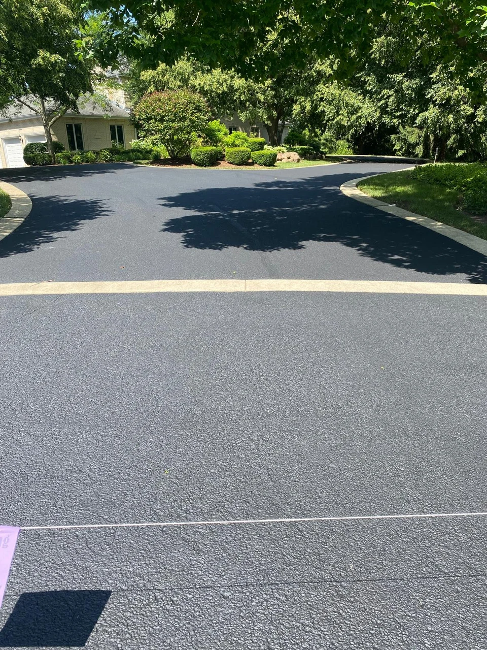 The same circular driveway after sealcoating, with dark asphalt, the same stone strip and planted island, and a cord across the entrance
